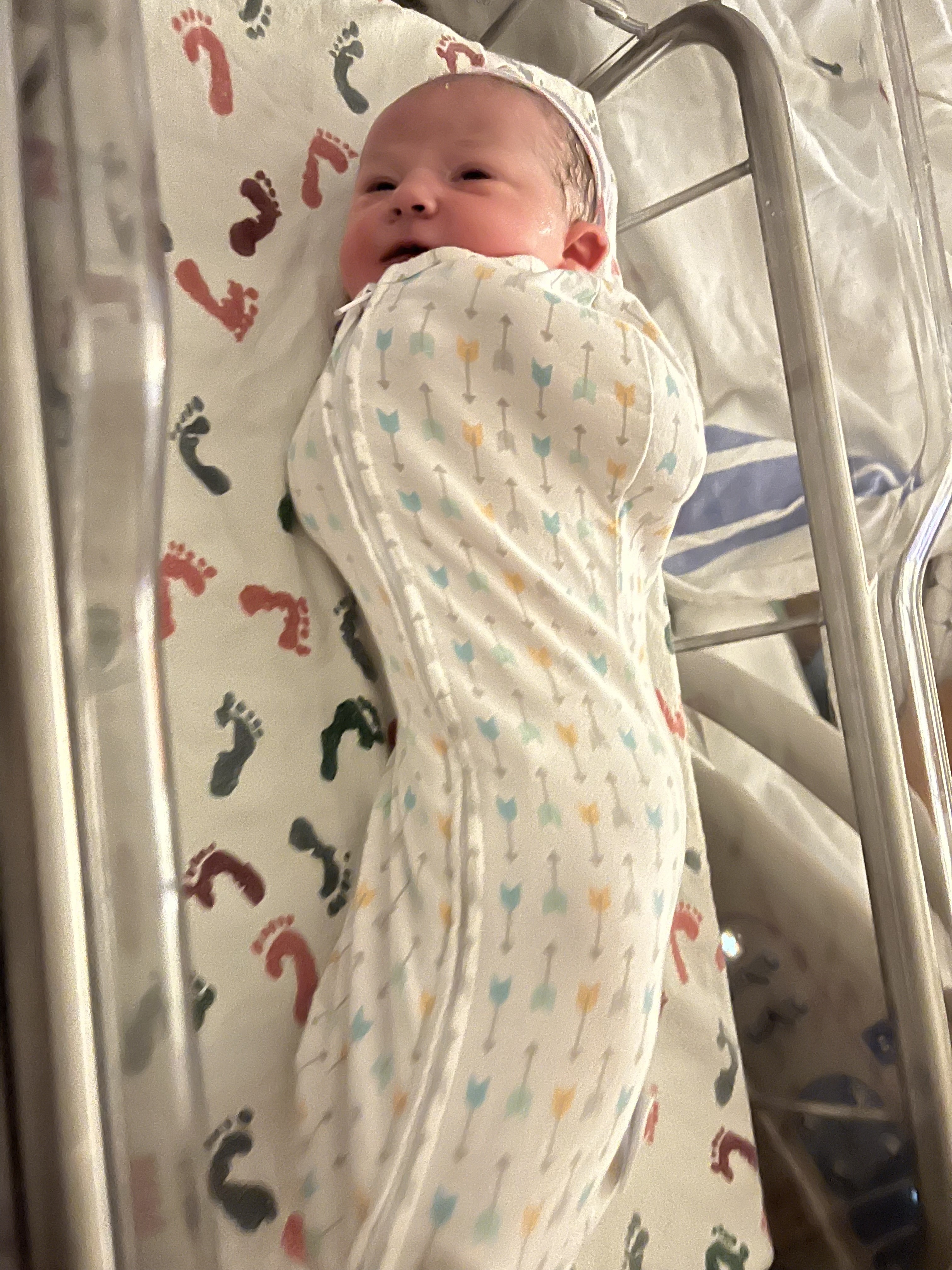 A newborn in a hospital nursery, swaddled and cozy. Their eyes are open, and there's blur along the bottom; their feet might be moving.